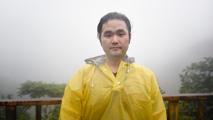 A man with a slightly resigned, discontent look on his face is standing in front of a fogged in viewpoint and he's wet and wearing a rain slicker. 