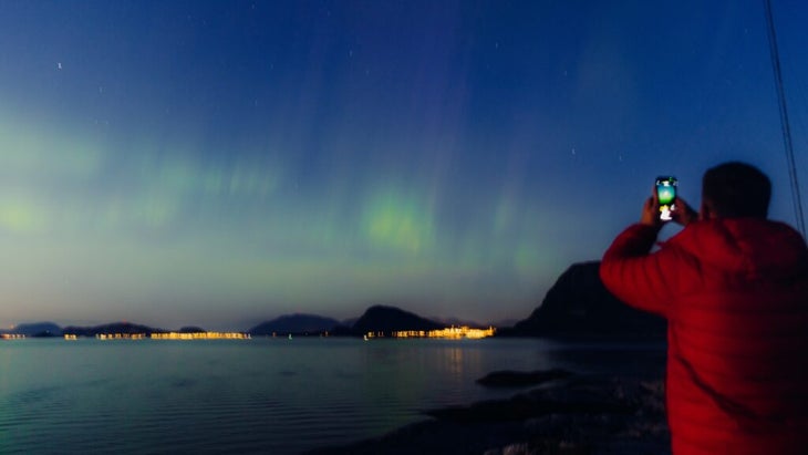 A man holds up his cell phone to shoot the northern lights illuminating the sky over a seascape near Alesund, Norway.