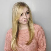 A portrait of Molly Longman. She has long blonde hair and is wearing a peach-colored sweater while standing in front of a grey wall.