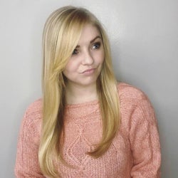 A portrait of Molly Longman. She has long blonde hair and is wearing a peach-colored sweater while standing in front of a grey wall.