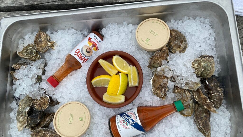 A tray of local sweet water oysters set amid ice, with a cup of lemons and some hot sauces and mignonette sauces placed within.