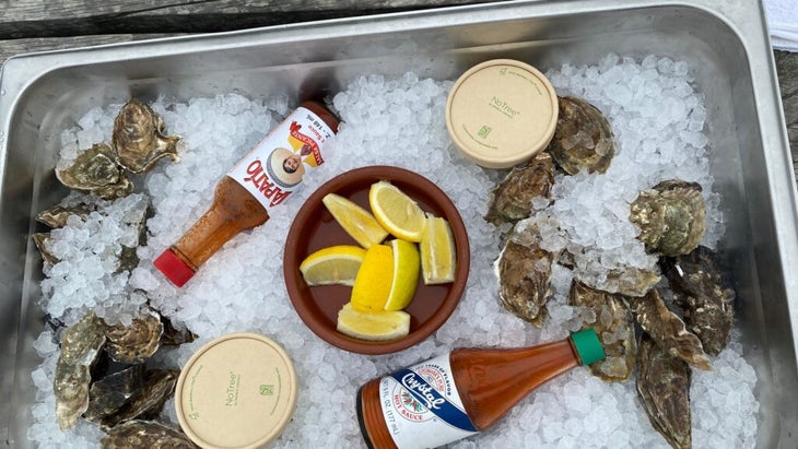 A tray of local sweet water oysters set amid ice, with a cup of lemons and some hot sauces and mignonette sauces placed within.