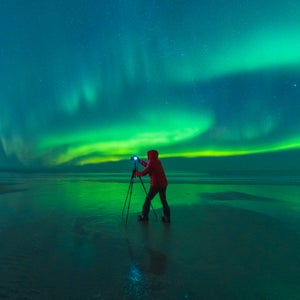 A man looks at the camera set up on his tripod as the green northern lights swirl on an Arctic horizon.
