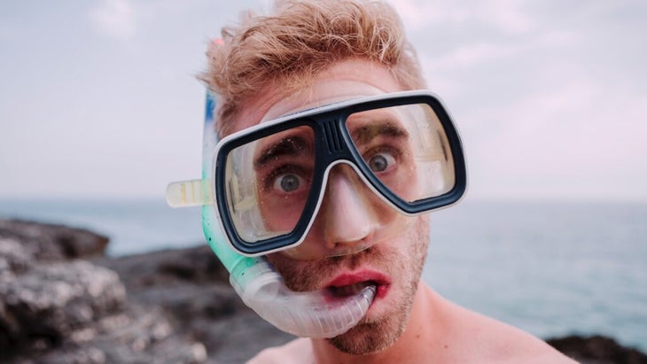 A man wearing a snorkel mask and tube standing in front of the ocean looks surprised and shocked.