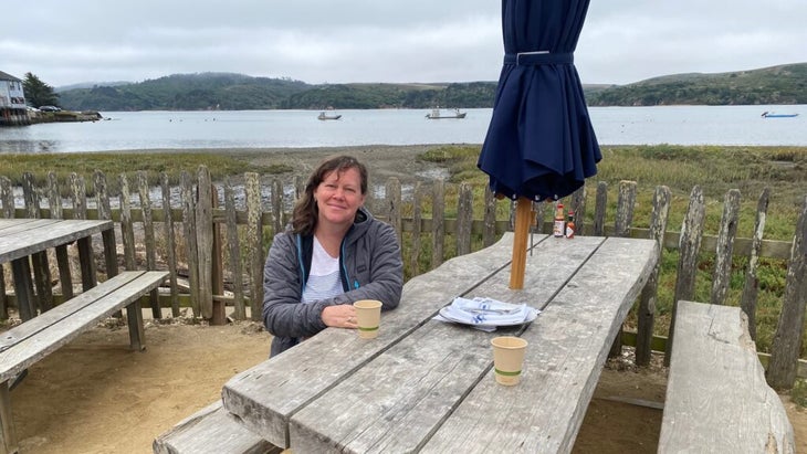 The author sits at a weathered picnic table, with Tomales Bay behind her and, farther in the distance, Point Reyes National Seashore.