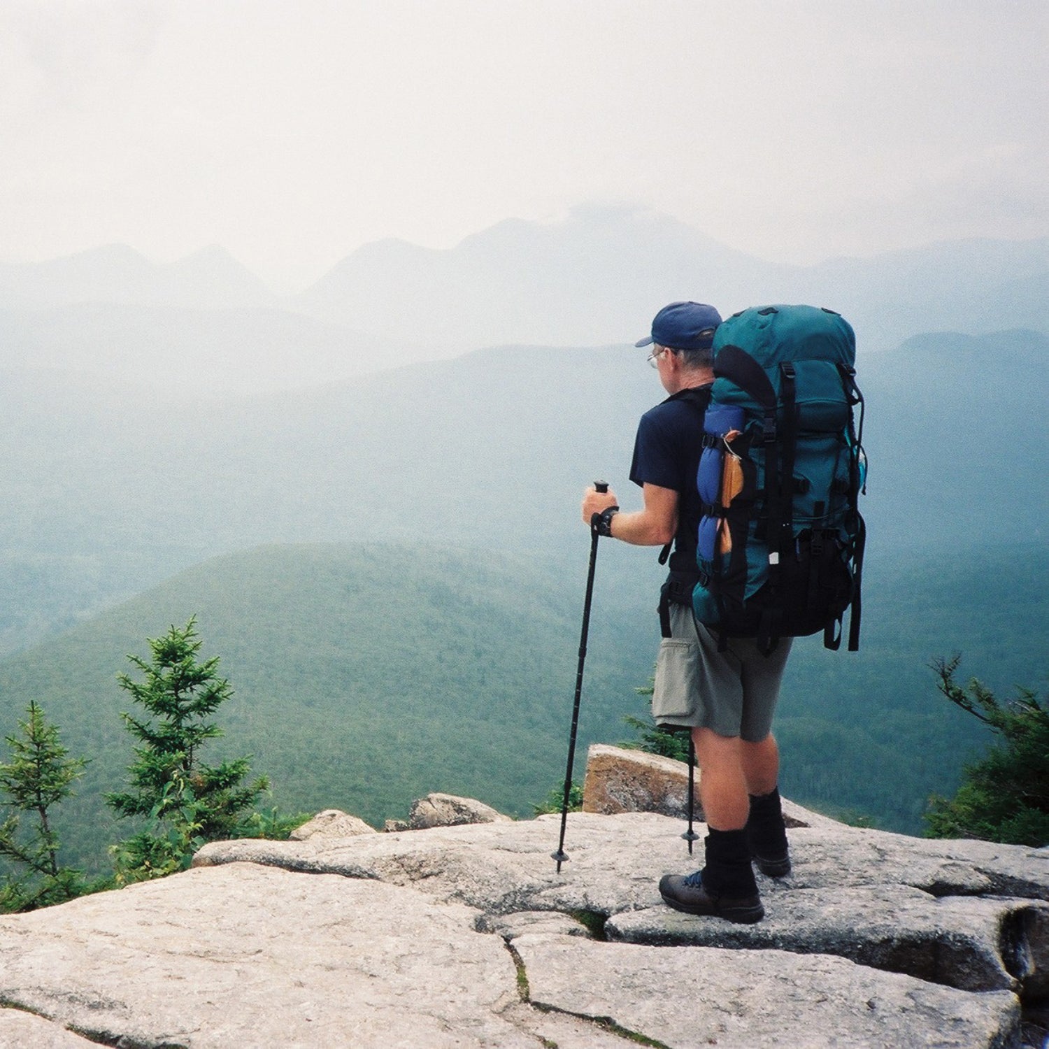 A middle-aged man wearing a large backpack and hiking boots looks over the edge of a rocky cliff toward mountains on a misty day.