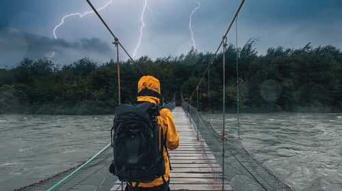 A person wearing a raincoat and a backpack looks across a river bridge they must cross, while three bolts of lightning are striking down the patch of land in front of them.
