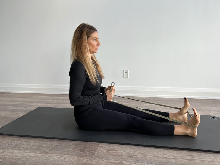 Woman sits on a mat with legs straight and resistance band pulled around the soles of her feet.