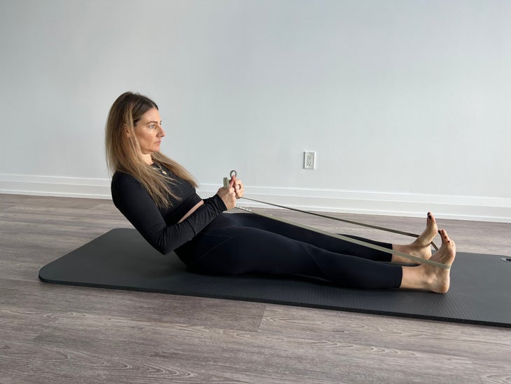 Woman sits on mat with legs extended in front of her and resistance band wrapped around the soles of her feet. She is holding the resistance band in her hands and leaning backwards.