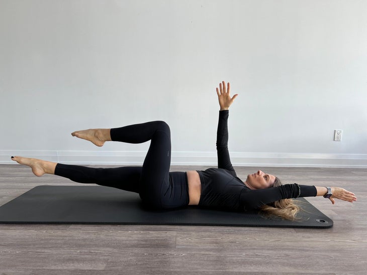 Woman lying on back with right arm straight into the air and left arm hovering straight above the floor. Left leg is bent and right leg is straight as both hover over the floor.