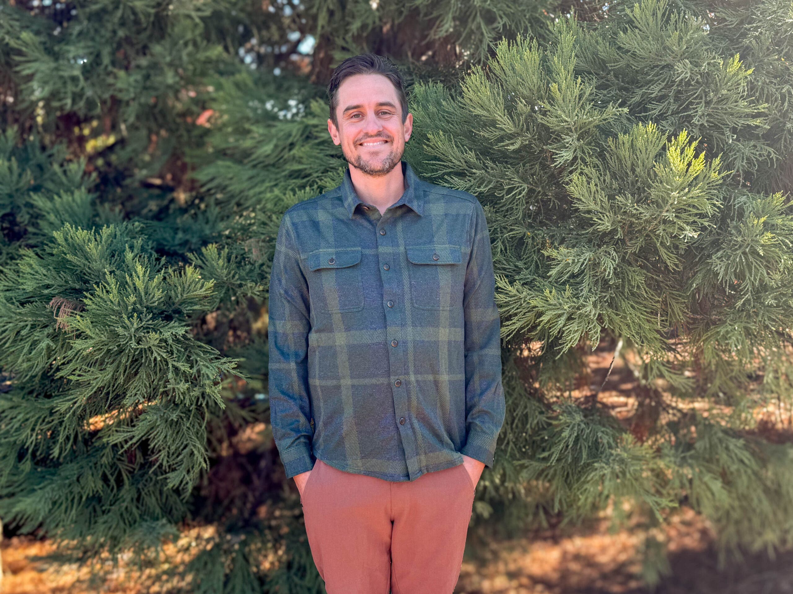 A man smiling while wearing the DUER Performance Flannel Button Down, one of the best flannel shirts for men for outdoor activities, featuring a green and navy plaid pattern and technical stretch fabric.