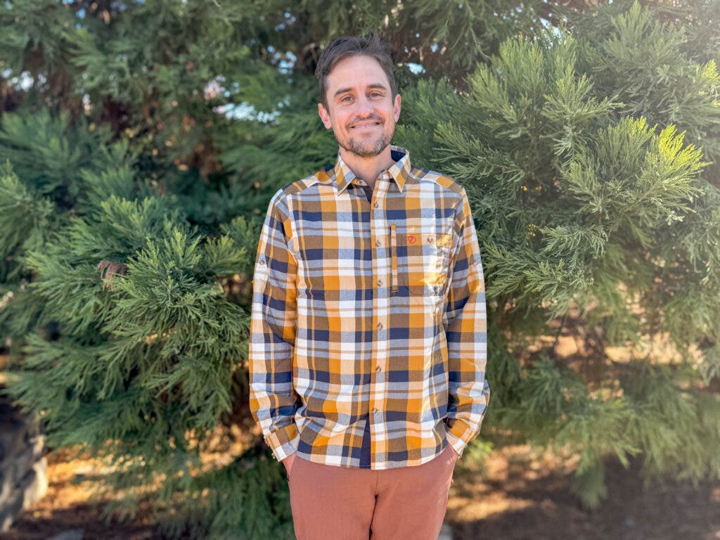 A man smiling outdoors wearing the Fjällräven Fjällglim Shirt in a gold, navy, and white plaid pattern, demonstrating its functional fit as one of the best flannel shirts for men for hiking and travel.