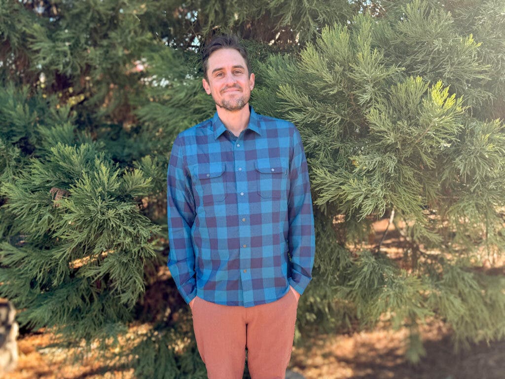 A man wearing the Flylow Handlebar Tech Flannel in a vibrant blue and navy buffalo check pattern, standing outdoors to showcase one of the best flannel shirts for men designed with technical performance in mind.