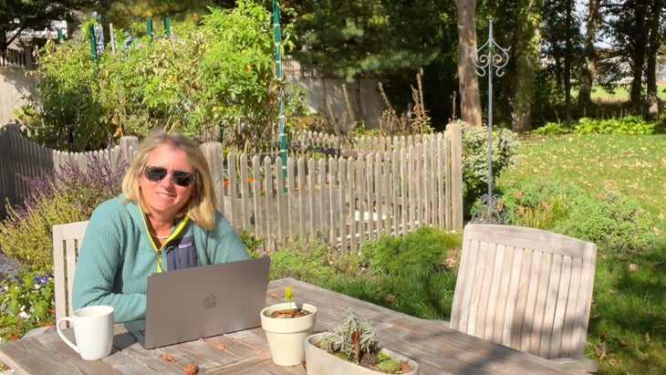 The author sitting in her garden at a table with coffee and her computer, enjoying the falling leaves.