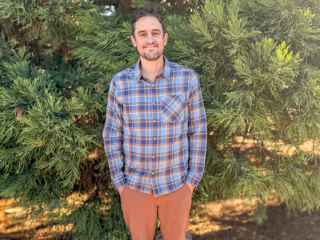 A man standing in front of evergreen trees wearing the Outdoor Research Ravenna Flannel, featuring a vibrant blue and orange plaid pattern, highlighted as one of the best flannel shirts for men for bright, outdoor style.