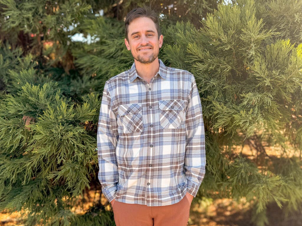 A man wearing the Royal Robbins Lost Coast Flannel in a blue, white, and brown plaid pattern, demonstrating why it is among the best flannel shirts for men for layering in wooded environments.