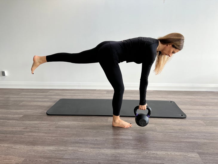 For one of the core exercises, a woman stands on her right leg leg while bending forward at the hip, with her left leg extended behind her. She is holding a kettlebell in her right hand.