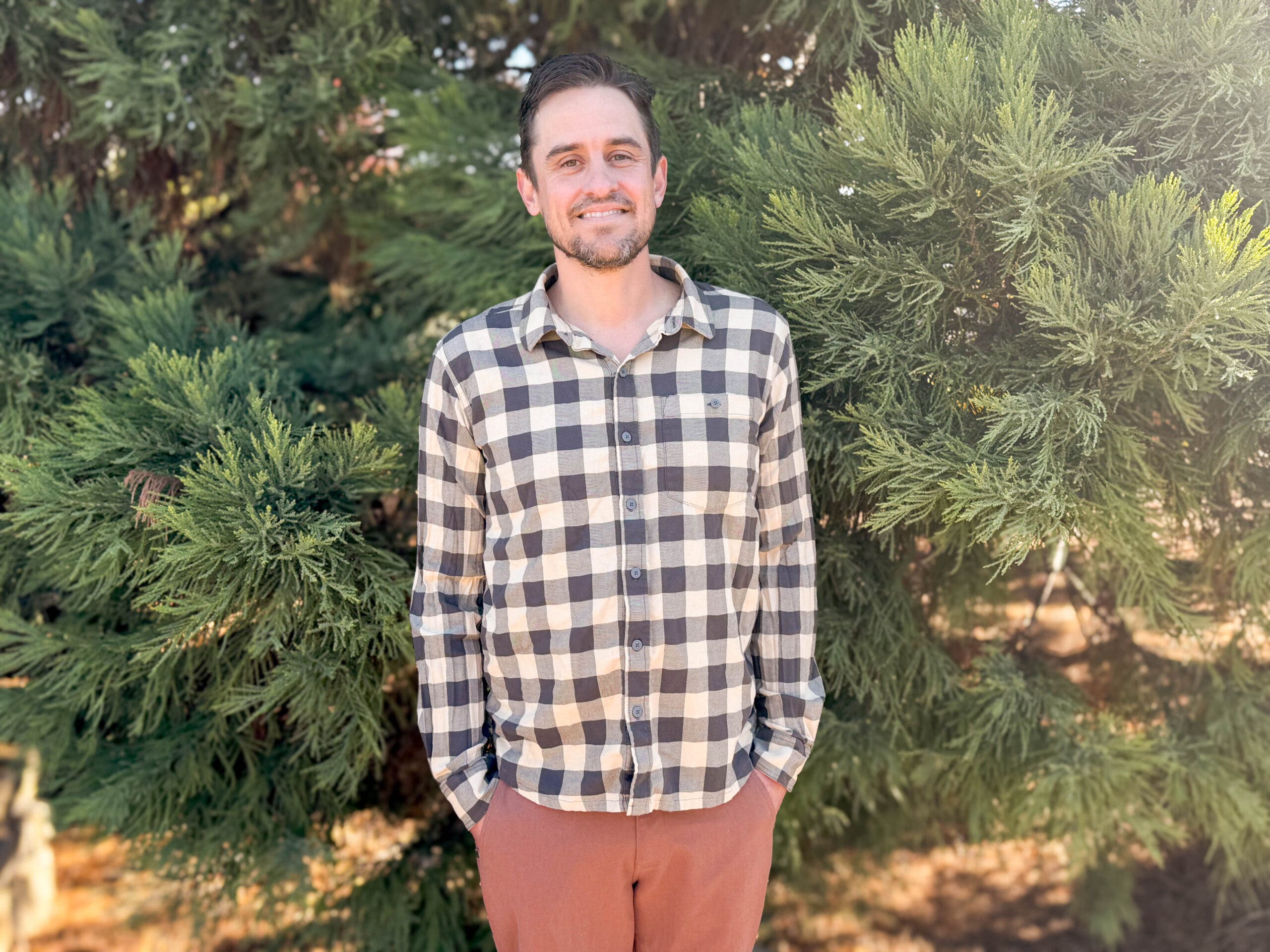 A man wearing the Stio Miter Stretch Lightweight Flannel in a cream and charcoal buffalo check pattern, standing against a backdrop of green foliage to highlight one of the best flannel shirts for men for transitional weather.