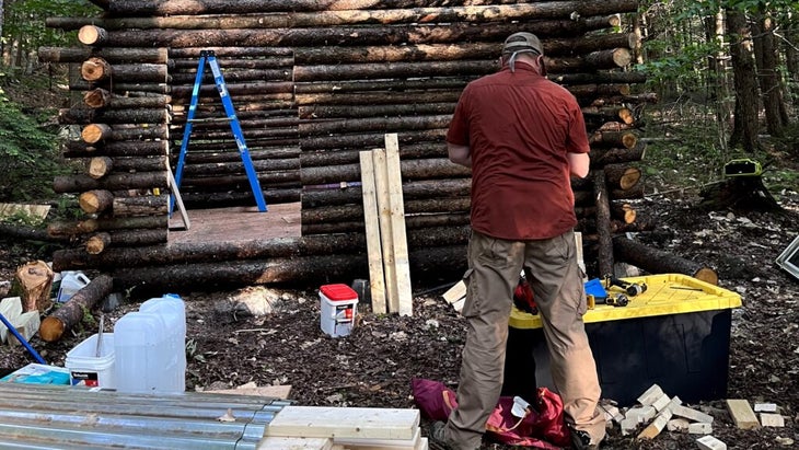 Josh Drinkard stands in front of a big plastic tub of tools in front of the log structure. 