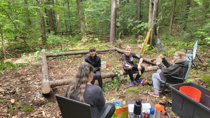 A mother sits next to their future cabin site with they three teens, eating in chairs. The first logs of the cabin are set up in a square behind them.