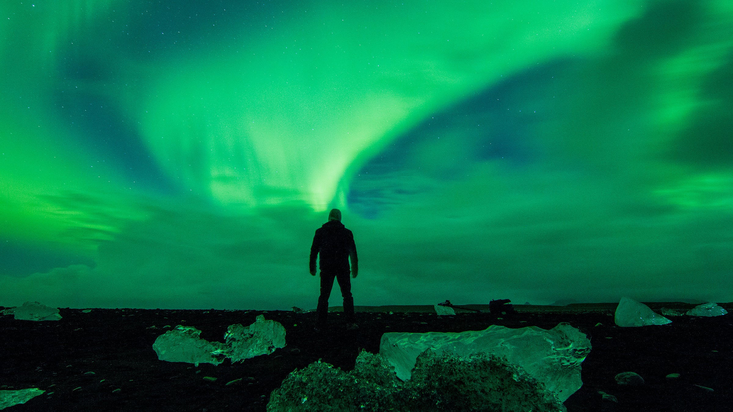 Man watching beautiful display of Aurora Borealis. Chunks of ice on black sand beach with dramatic sky. Majestic view of nature at night.