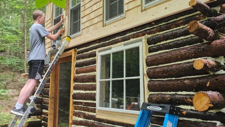 Josh Drinkard’s teenage son, Mason, attaches shingles to the second level exterior, working from a ladder leaning against the structure.