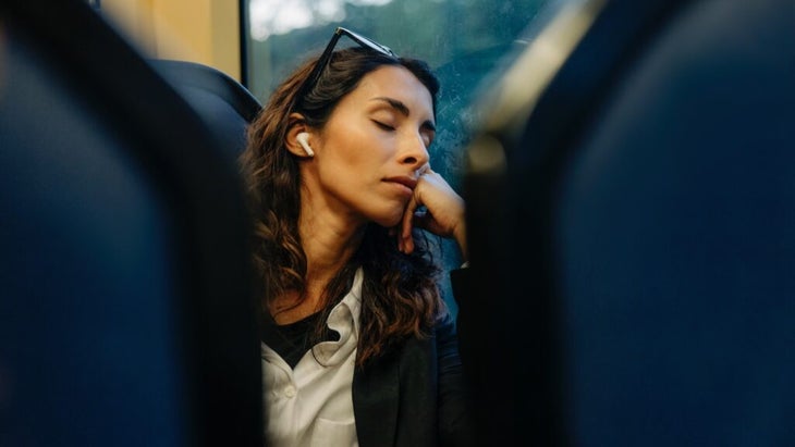 A woman wearing earpods sleeps on what appears to be a busy, with her head tilted toward the window.