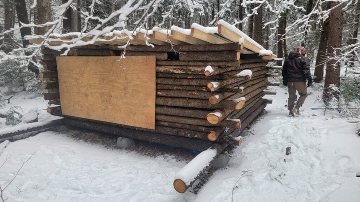 The lower half of the cabin is covered with a makeshift roof and plywood sheet nailed over the door, with a few inches of snow covering the structure and ground.