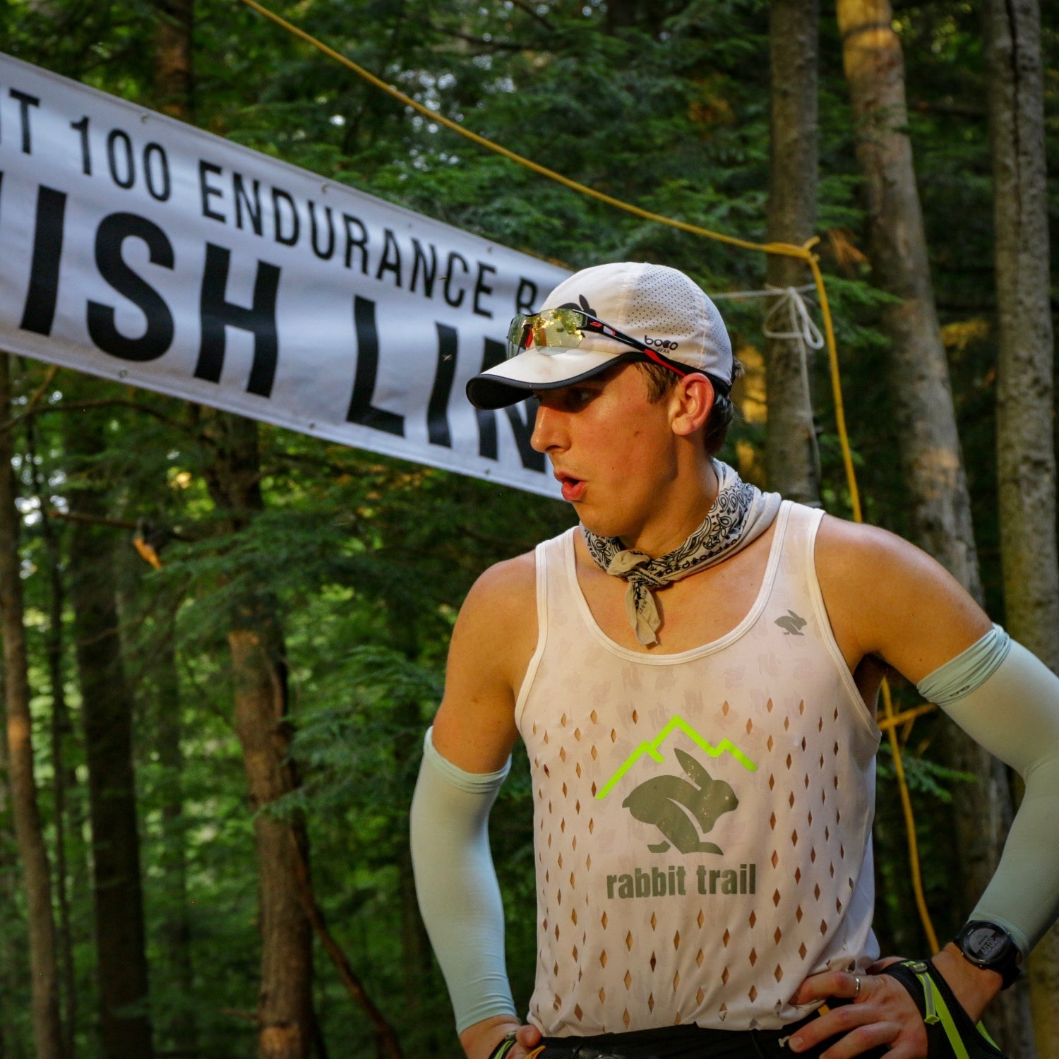 A man in compression sleeves, a tank top, and a baseball cap exhales at the finish line of the Vermont 100 endurance race