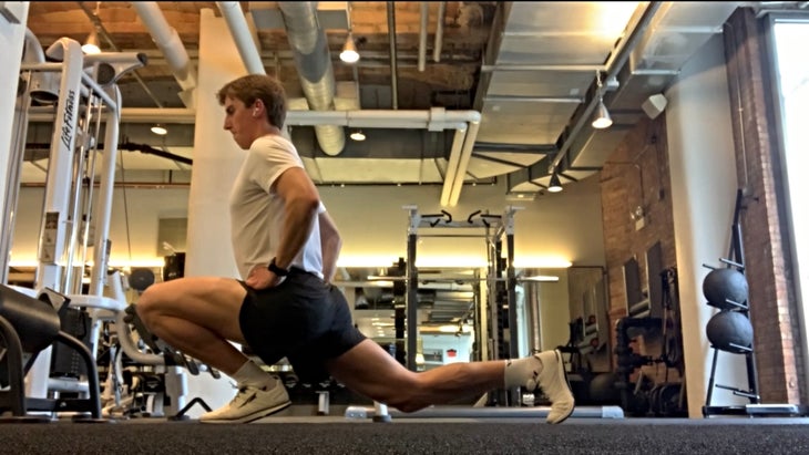 A young man lunges in a gym weight room n a white t-shirt and black shorts, with his knee far beyond his toes 