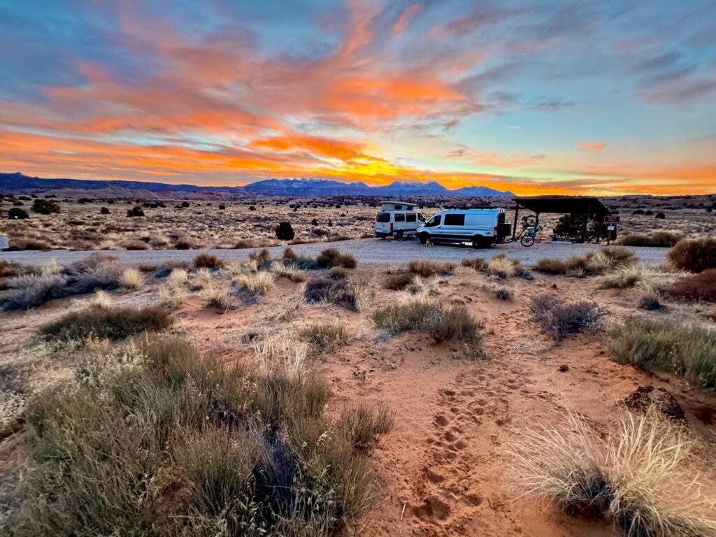 A spectacular sunset shows clouds colored in peach and yellow. Two camper vans are parked at a campsite near Moab, Utah.
