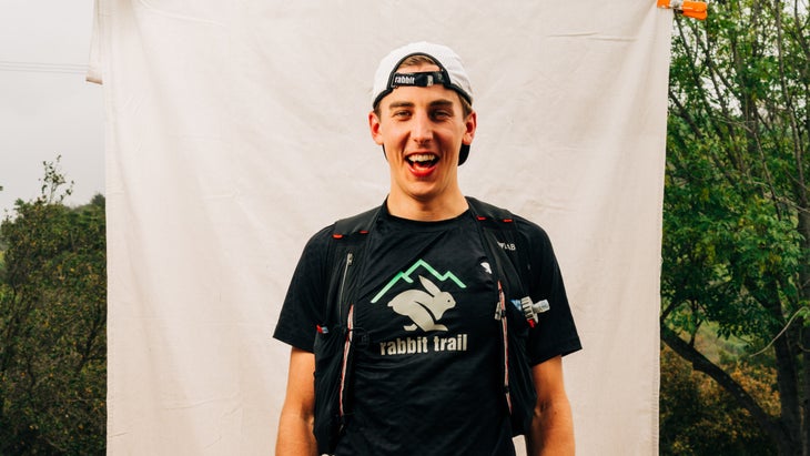 A man in a backwards baseball cap smiles for the camera while wearing a black Rabbit-branded running t-shirt and a running vest