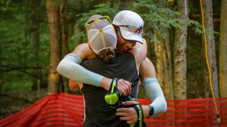 A man in a baseball cap and compression sleeves holding water bottles in each hands hugs a woman in a baseball cap. They are outdoors in the forest.