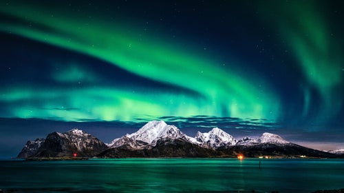 A huge, swirling green aurora illuminates the night sky over snowcapped mountains and the sea in Greenland