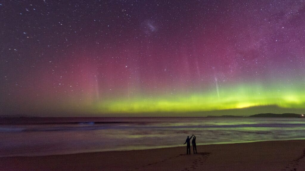 A silhouette of a couple on the shore of Tasmania while bioluminescent waves roll in and the southern lights shine on the horizon.