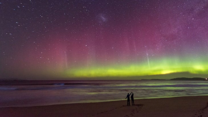 A silhouette of a couple on the shore of Tasmania while bioluminescent waves roll in and the southern lights shine on the horizon.