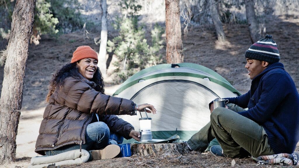 A man and woman wearing beanies and puffy jackets sit in from of their tent.