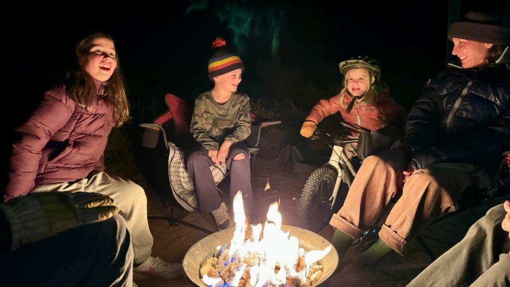 A group of kids—one on a small bike—gather around a campfire at night.