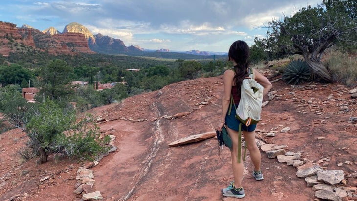 A woman wearing a backpack and trail-running shoes looks out over the high-desert landscape above the Enchantment Resort to greater Sedona.