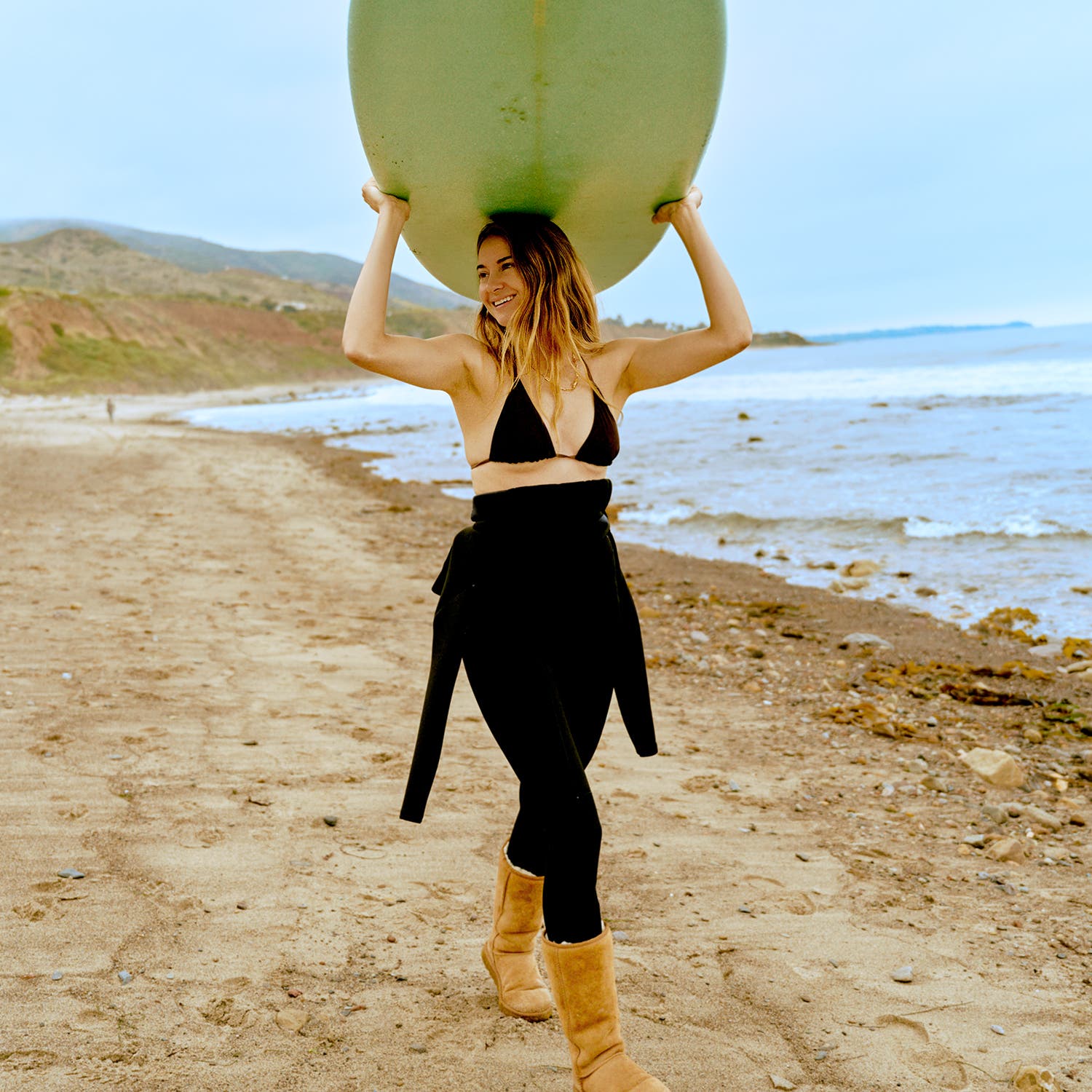 Shailene Woodley walks with a surfboard on the beach. Wetsuit by Patagonia, boots by Ugg, bikini top from Araks