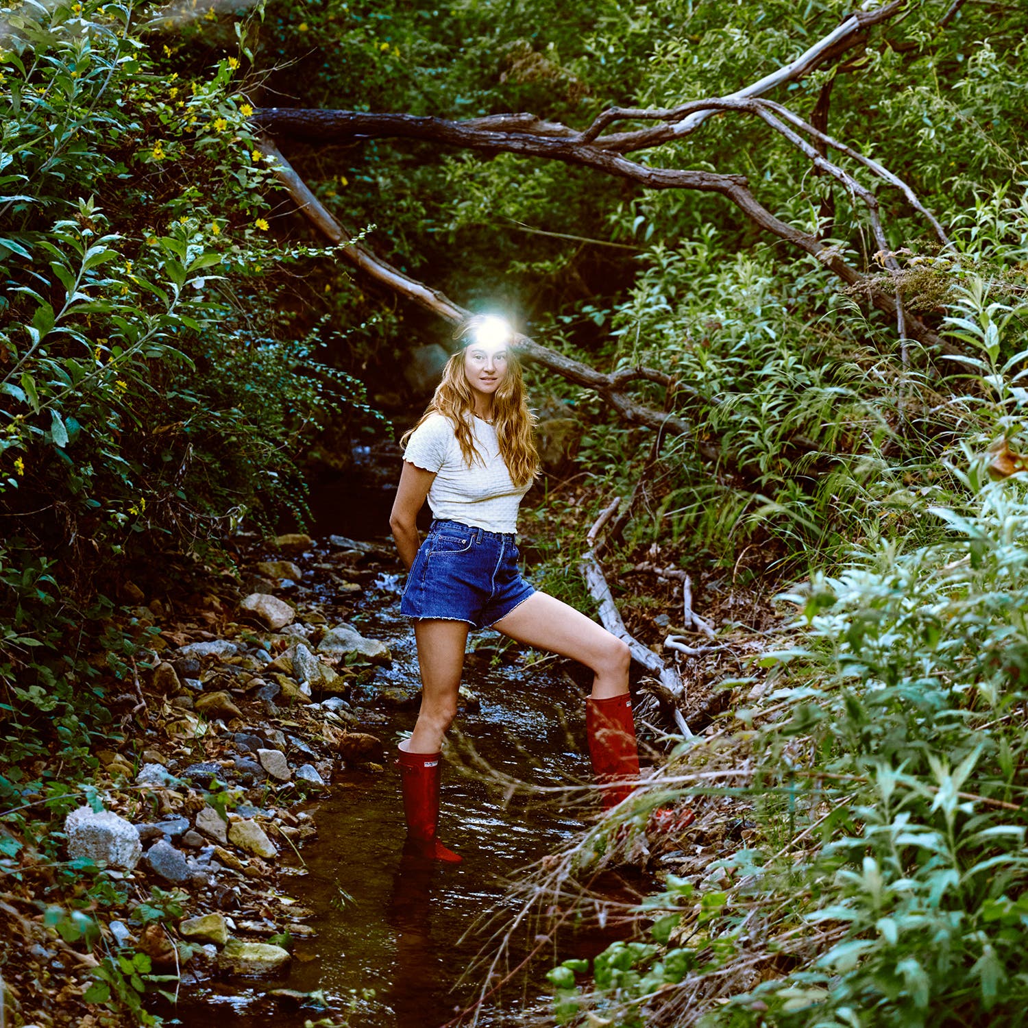 Shailene Woodley with a headlamp, standing in a creek. Checker Baby Tee by Guest in Residence, Denim Shorts by AGOLDE, Original Tall Rain boots by Hunter, jewelry by Handle Only with Love