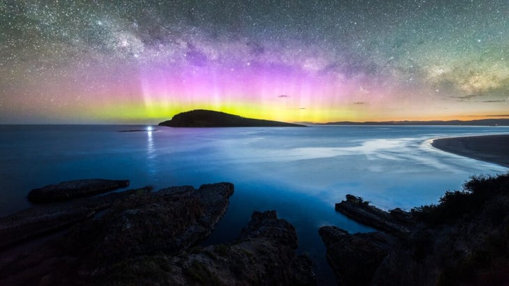 The southern lights shine over a silhouetted seascape above Tasmania, Australia.