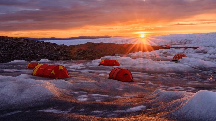 Three red tents pitched on the Greenland ice sheet glow in the fading light of the sun.