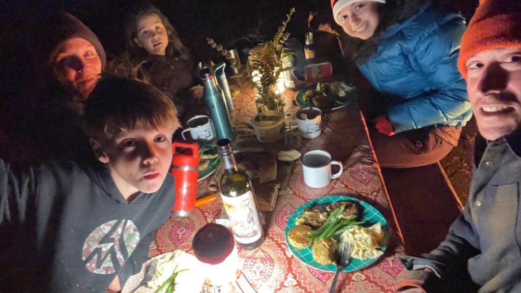A family enjoys their Thanksgiving dinner—pasta, crab cakes, and green beans—at a picnic table, lit by lanterns.