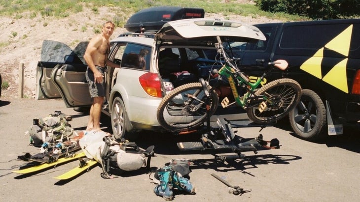 A man standing near the open passenger door of his car, with the trunk popped, and more than a dozen items of outdoor gear surrounding the car, including a mountain bike. 