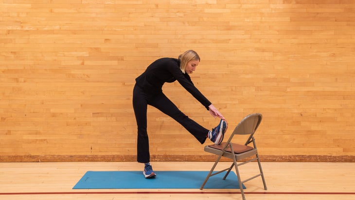 A woman in a gym puts her left leg on top of a chair and bends forward at the waist to stretch toward her foot, grabbing her foot with her hands.