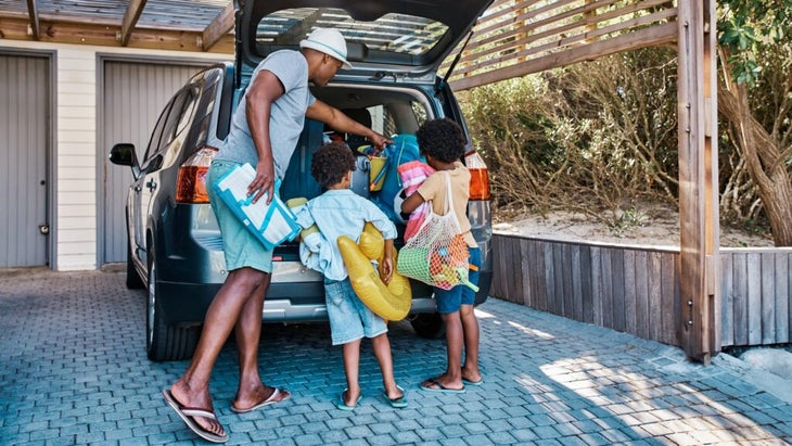 A father packing a car is helped by his two young children, whose hands are full of vacation items. Dad points where to put them in the trunk.