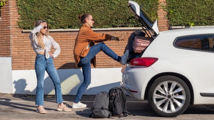 One woman kicks a suitcase into a car trunk, in an attempt to make it fit, while another woman looks on, frustrated. 