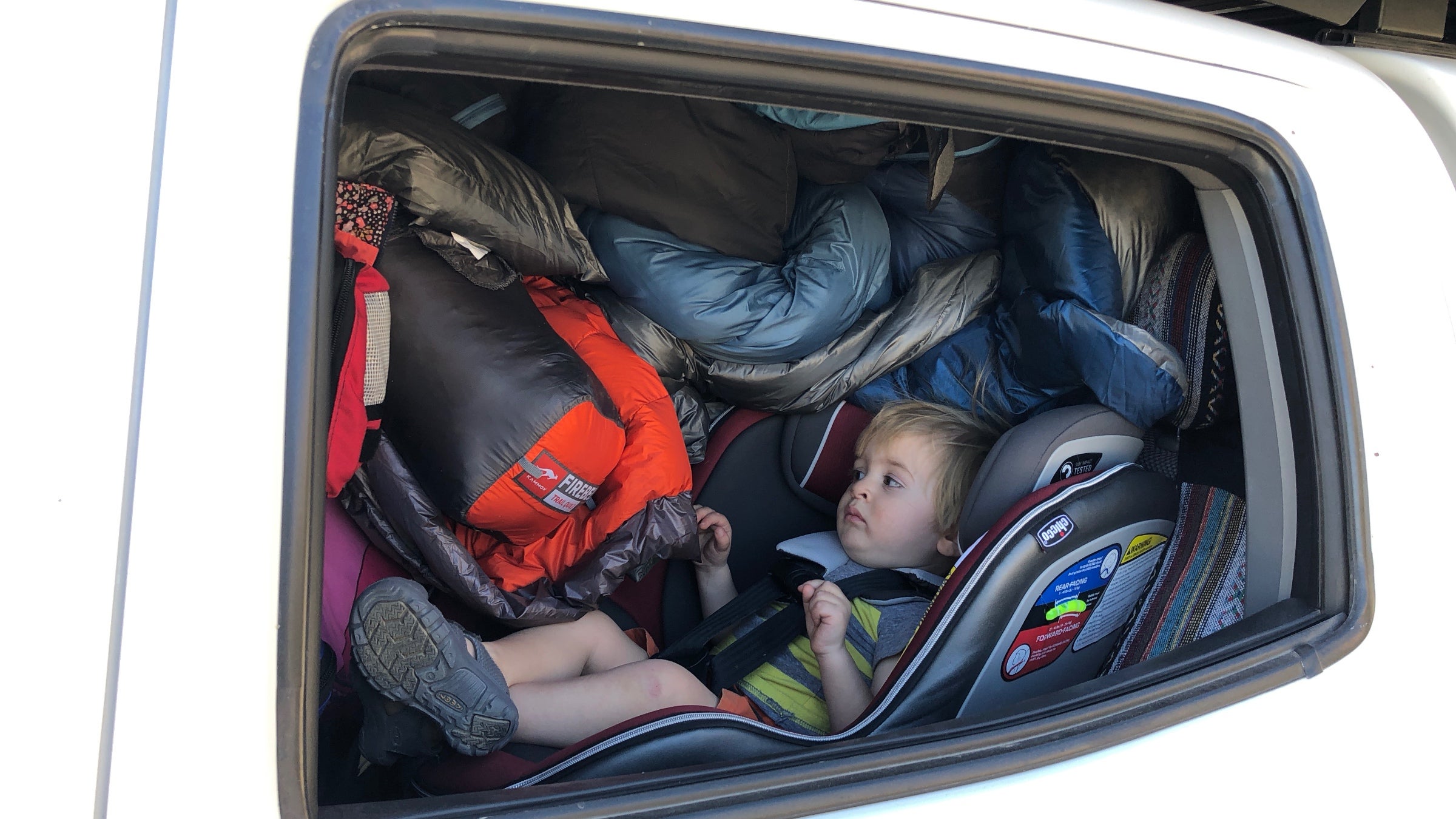 A toddler in his car seat is wedged next to the window and the entire space to the right of him is full of sleeping bags, packs, and other gear.