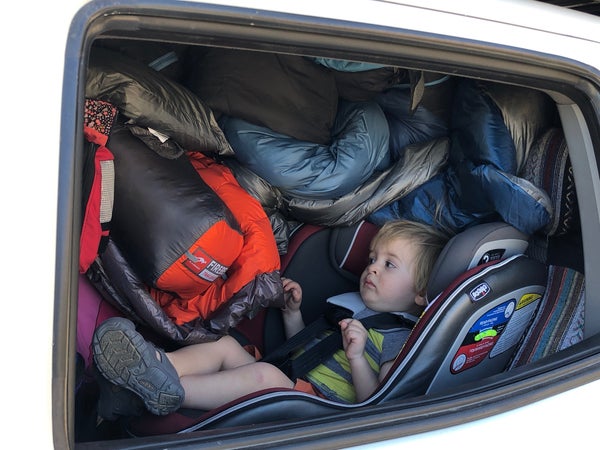 A toddler in his car seat is wedged next to the window and the entire space to the right of him is full of sleeping bags, packs, and other gear.